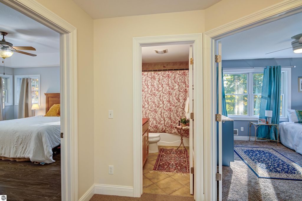 Interior view of a Traverse City home, showcasing a hallway with doorways leading to a bedroom, bathroom with floral shower curtain, and another bedroom featuring large windows and decorative curtains.