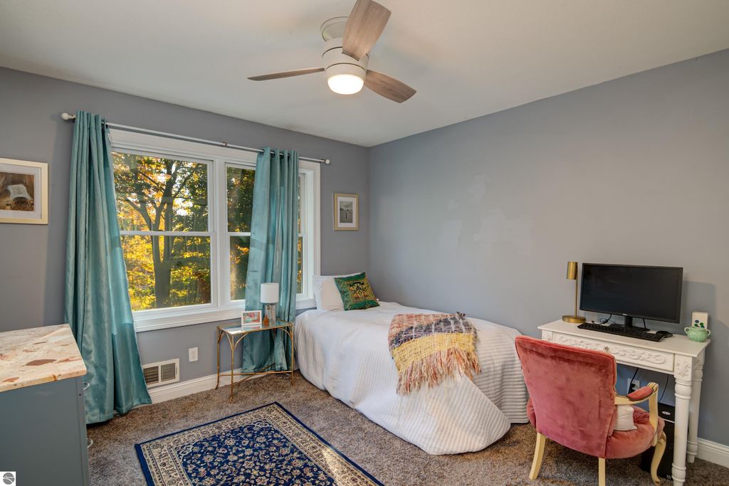 Cozy bedroom in Traverse City home featuring a bed with a decorative blanket, a window with scenic views, teal curtains, a small desk with a TV, and a plush pink chair, showcasing modern comfort and inviting design.