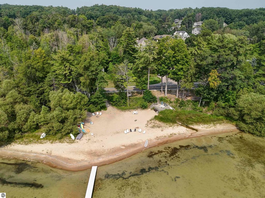 Aerial view of beach area with sandy shore, surrounded by trees and homes, featuring lounge chairs, kayaks, and a dock on East Grand Traverse Bay near 6436 Peregrin Court, Traverse City, MI.