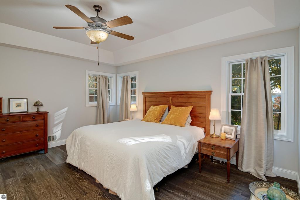 Brightly lit bedroom featuring a wooden bed with yellow accent pillows, hardwood flooring, and large windows with curtains, showcasing a cozy and inviting atmosphere in the Traverse City home for sale.