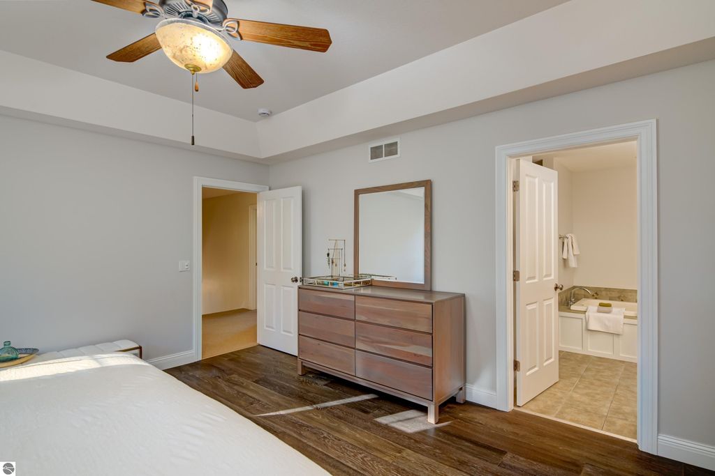 Master bedroom interior featuring a wooden dresser with a mirror, ceiling fan, and adjoining bathroom entrance, showcasing modern design and hardwood flooring.