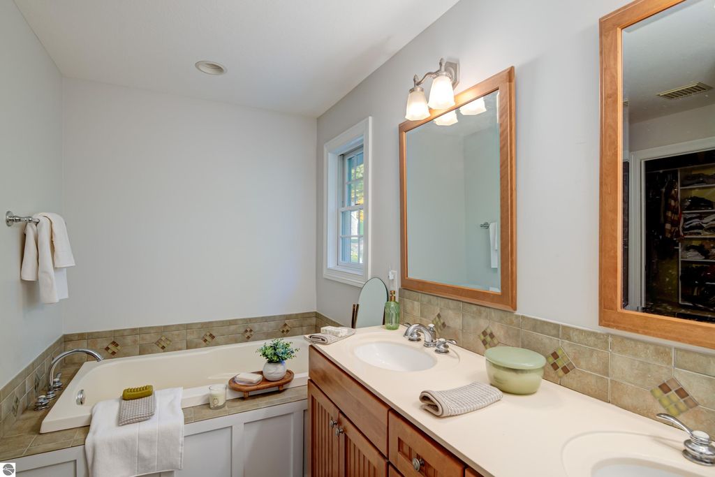 Luxurious bathroom featuring a double vanity with wooden cabinetry, a soaking tub, decorative tile accents, and natural light from a window, enhancing the modern comfort of the home at 6436 Peregrin Court, Traverse City, MI.
