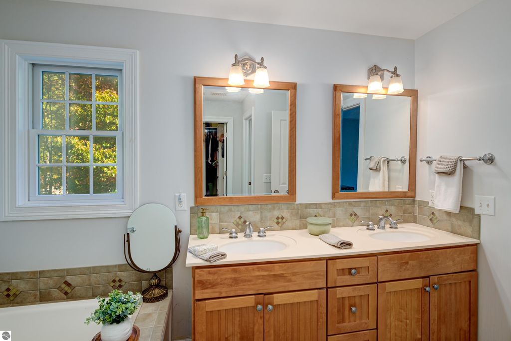 Bathroom with dual sinks, wooden cabinetry, large mirrors, and a window, featuring decorative tile accents and a plant on the counter, showcasing a bright and inviting atmosphere.