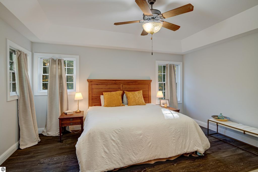 Cozy bedroom featuring a wooden bed frame with decorative pillows, bedside lamps, and large windows with curtains, showcasing a warm and inviting atmosphere in the Traverse City home for sale at 6436 Peregrin Court.