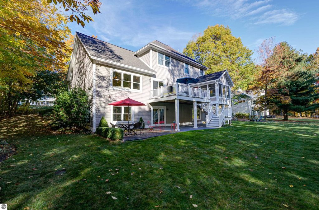 Custom-designed home exterior at 6436 Peregrin Court, Traverse City, featuring a landscaped yard, patio with seating and umbrella, and a walkout lower level, surrounded by trees in autumn colors.