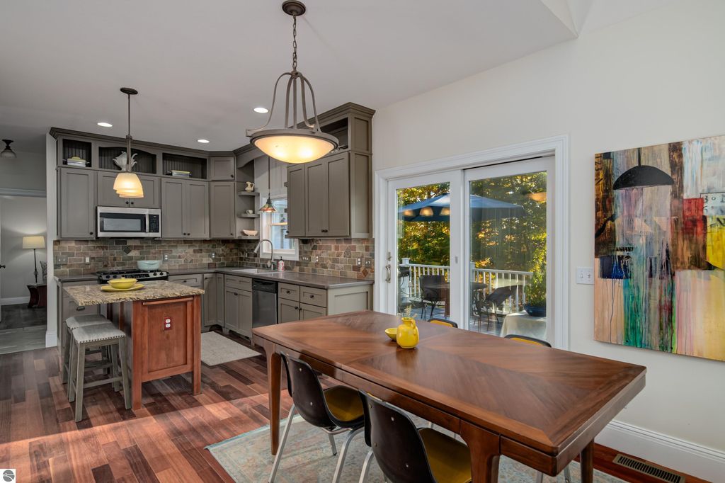 Modern kitchen with gray cabinetry, stainless steel appliances, and a wooden dining table set for four, featuring a yellow centerpiece, and large windows overlooking a deck with outdoor seating.