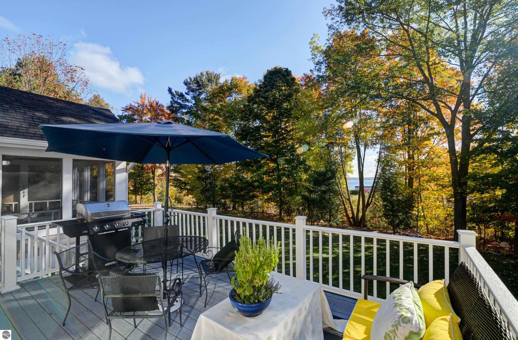 Deck area with outdoor dining set, grill, and umbrella, surrounded by trees showcasing autumn colors, with distant view of East Grand Traverse Bay, emphasizing the property's outdoor living space at 6436 Peregrin Court, Traverse City, MI.