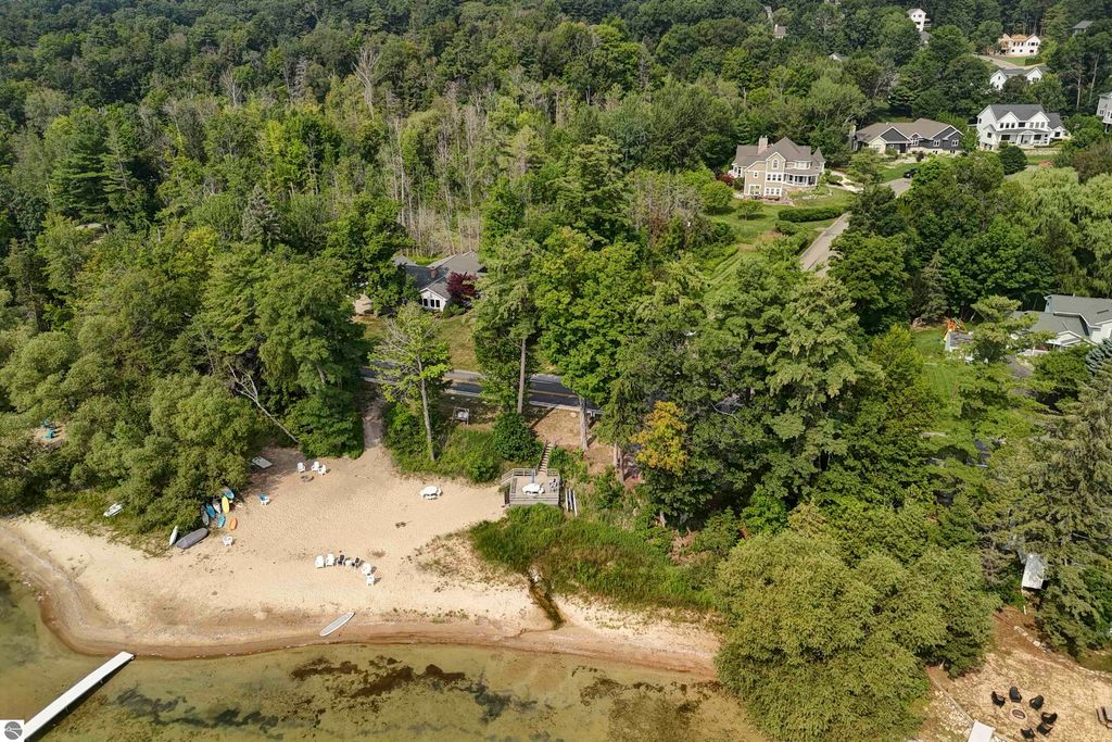 Aerial view of 6436 Peregrin Court property in Traverse City, showcasing sandy beach, East Grand Traverse Bay, surrounding trees, and nearby homes.