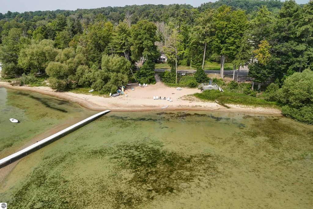 Aerial view of a sandy beach with shared frontage on East Grand Traverse Bay, featuring a wooden dock, lush greenery, and a park-like setting near 6436 Peregrin Court, Traverse City, MI.