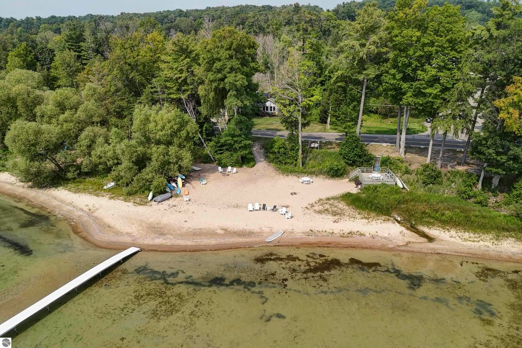 Aerial view of a sandy beach with shared frontage on East Grand Traverse Bay, featuring lounge chairs, kayaks, and a dock, surrounded by lush trees near 6436 Peregrin Court, Traverse City, MI.