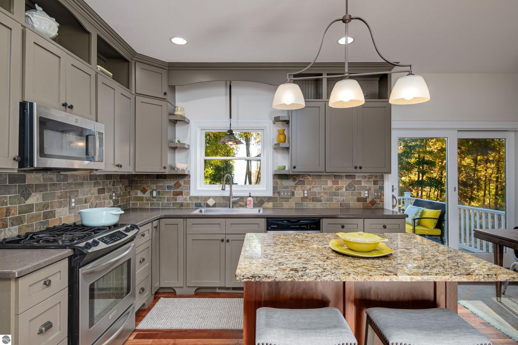 Modern kitchen featuring granite countertops, stainless steel appliances, and a spacious island with yellow plates, complemented by natural light from large windows overlooking a landscaped yard.