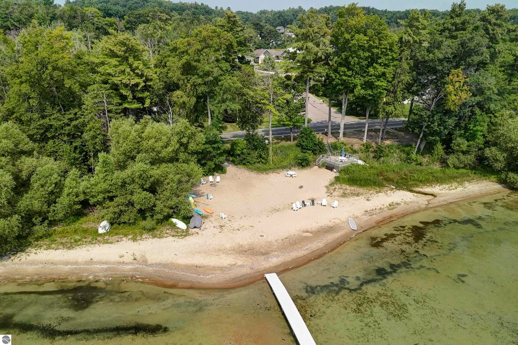 Aerial view of a sandy beach with chairs and kayaks, surrounded by lush trees and a clear shoreline on East Grand Traverse Bay near 6436 Peregrin Court, Traverse City, MI.