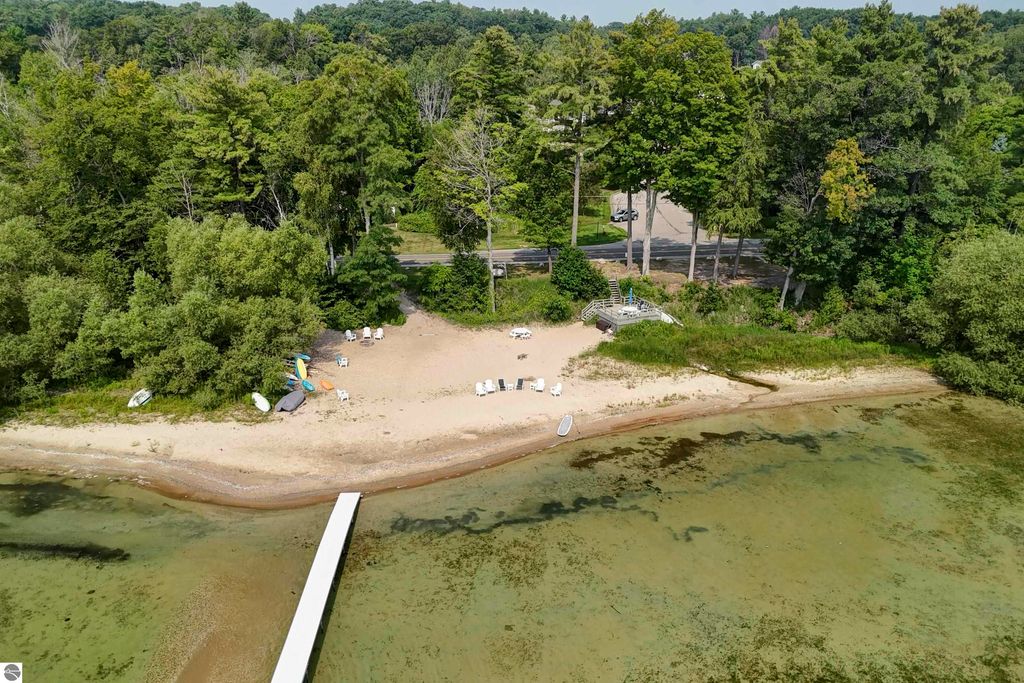 Aerial view of a sandy beach with shared frontage on East Grand Traverse Bay, featuring lounge chairs, kayaks, and a wooden dock, surrounded by lush greenery and trees near 6436 Peregrin Court, Traverse City, MI.