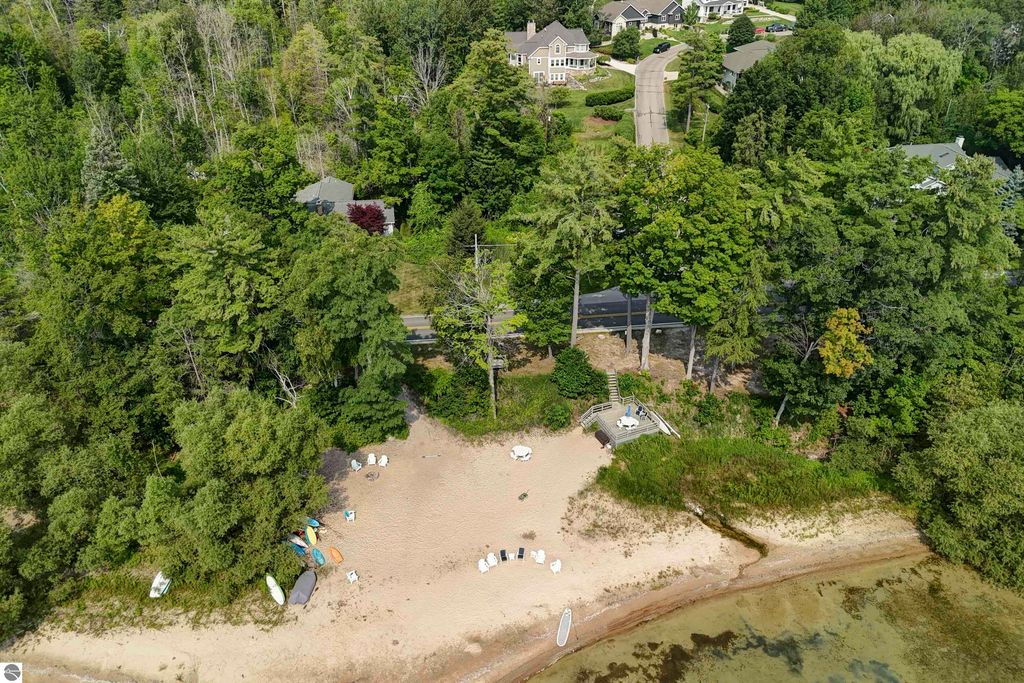 Aerial view of a sandy beach with shared frontage on East Grand Traverse Bay, surrounded by lush greenery and residential homes, featuring beach chairs and kayaks, highlighting the scenic location of 6436 Peregrin Court, Traverse City, MI.