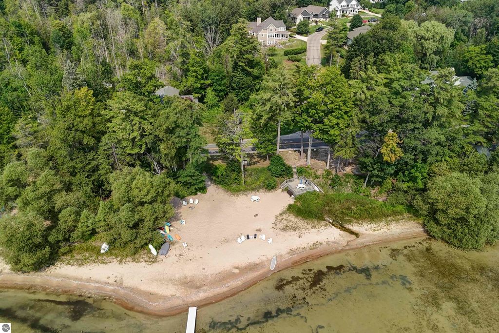 Aerial view of a sandy beach with shared frontage on East Grand Traverse Bay, surrounded by lush greenery and residential homes, featuring beach chairs and watercraft.