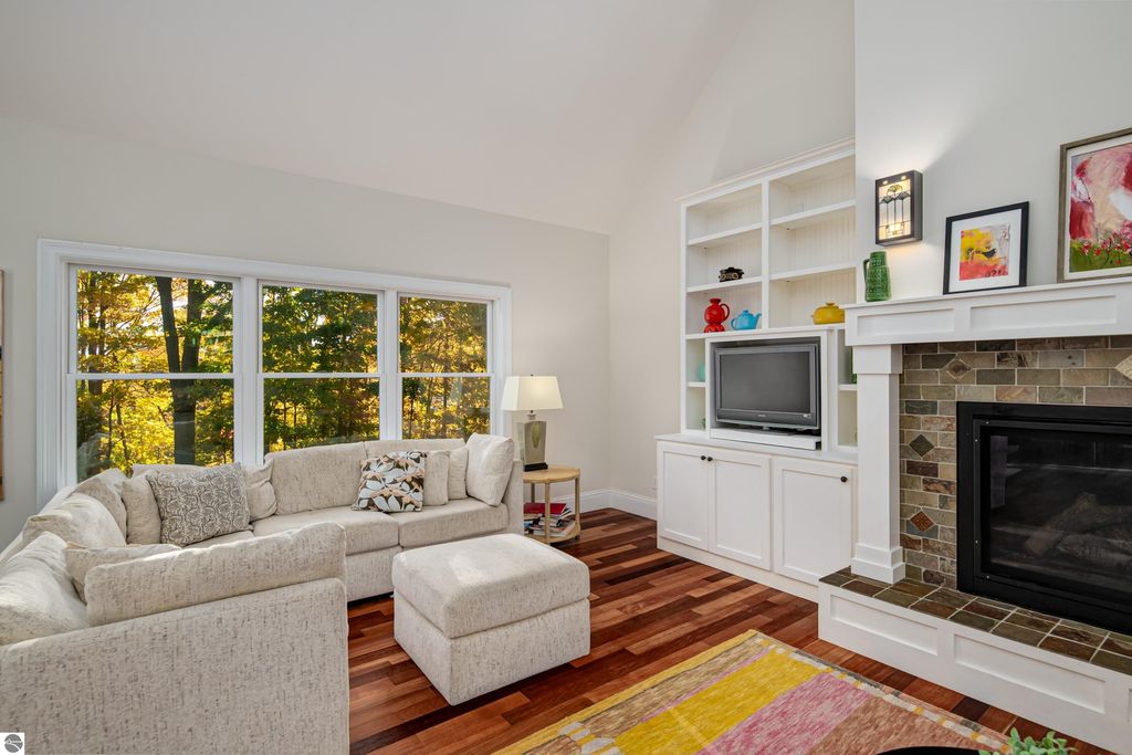 Cozy great room with a sectional sofa, gas fireplace, and large windows showcasing autumn views, highlighting the custom design and inviting atmosphere of the home at 6436 Peregrin Court, Traverse City, MI.