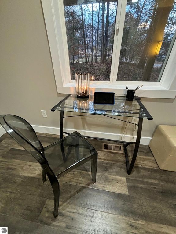 Modern workspace with a glass desk, transparent chair, tablet, and decorative candle, featuring a view of trees through a window, enhancing the home's comfort and convenience.
