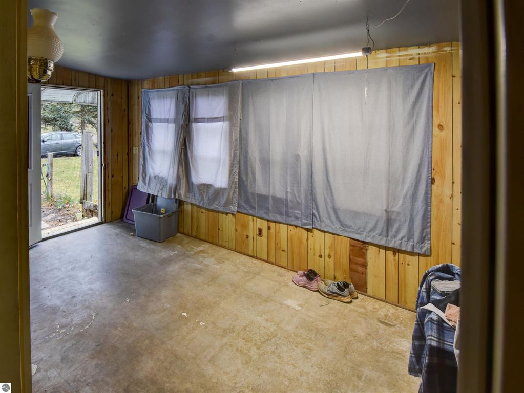 Interior view of a home entryway at 974 Parkway Drive, Tawas City, featuring wooden paneling, gray curtains, and a cement floor, with a door leading to the yard and shoes on the floor.
