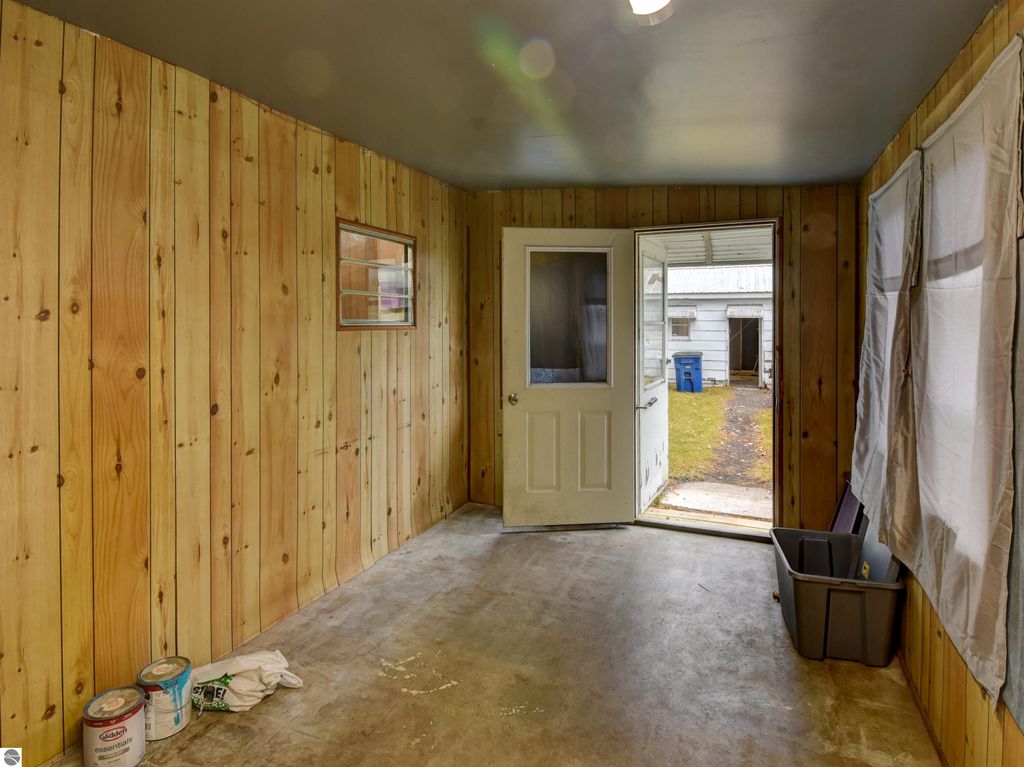 Interior view of a garage with wooden paneling, cement floor, open door leading outside, and storage items, highlighting the potential for hobbies and projects in Tawas City, MI property listing.