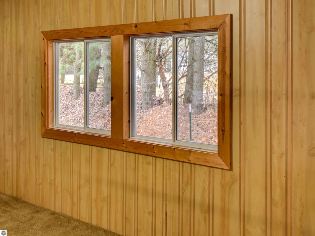 Interior view of a room featuring a large window with wooden trim, showcasing a view of trees outside, against a backdrop of wooden paneling and carpeted flooring, relevant to the real estate listing for a home at 974 Parkway Drive, Tawas City, MI.