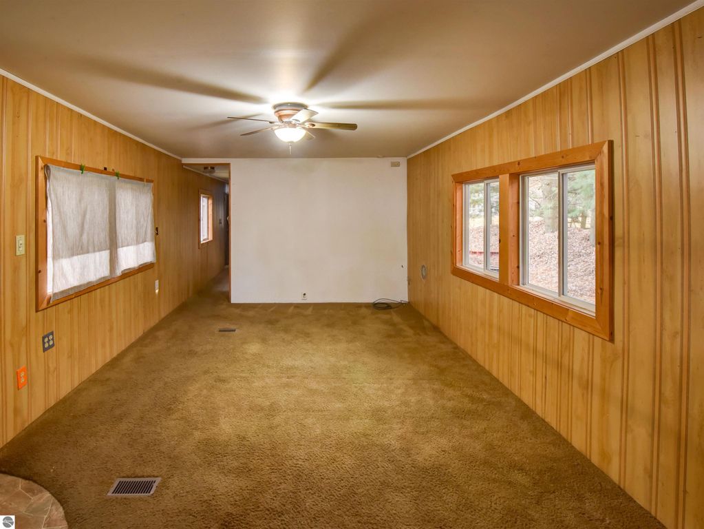 Interior view of a cozy living room in a Tawas City home, featuring wood paneling, carpeted flooring, and multiple windows allowing natural light, ideal for a starter house.