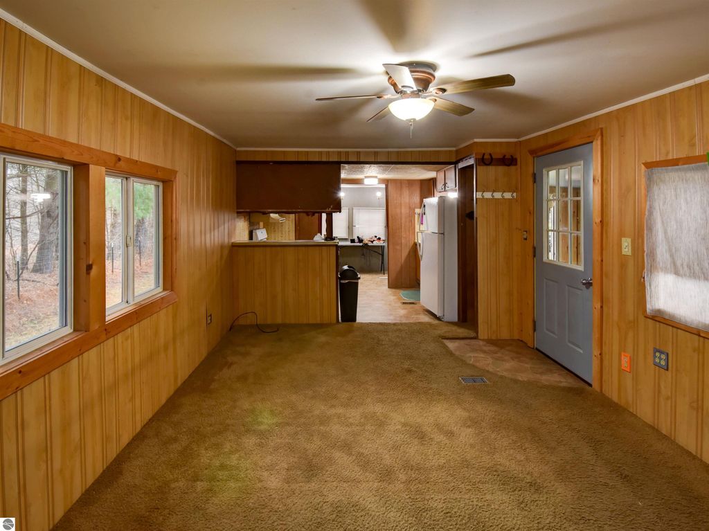 Interior view of a cozy living space in a Tawas City home, featuring wooden paneling, carpeted flooring, a ceiling fan, and a kitchen area visible in the background.