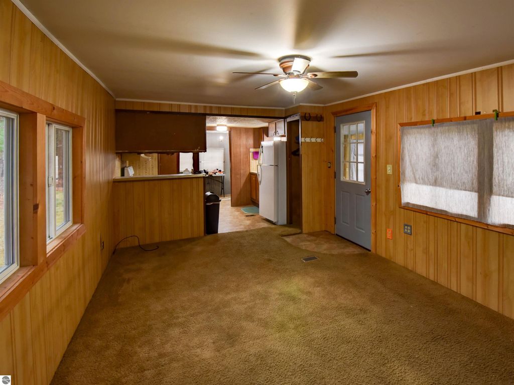 Interior view of a cozy living space in a 2-bedroom home, featuring wood paneling, a ceiling fan, and a kitchen area with a refrigerator, ideal for a starter house in Tawas City, MI.