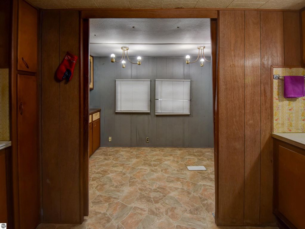 Interior view of a cozy kitchen space at 974 Parkway Drive, Tawas City, featuring wooden cabinetry, a tiled floor, and a view into a well-lit room with two windows and decorative light fixtures, highlighting the home's potential for updates.