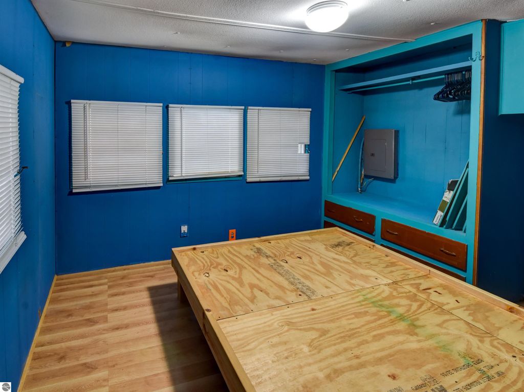 Interior of a blue-walled bedroom featuring a wooden platform bed, three windows with blinds, and a closet with shelves and storage drawers, suitable for a starter home in Tawas City, MI.