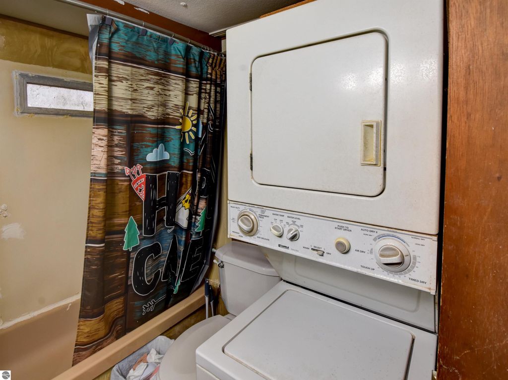 Laundry area with stacked washer and dryer, colorful "Happy Camper" shower curtain, and window, showcasing a cozy home feature in Tawas City, MI real estate listing.