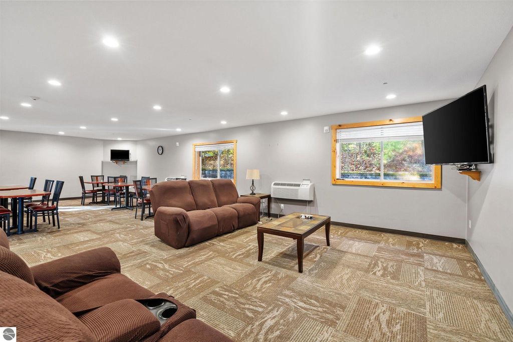 Interior view of a cozy lounge area in a motel, featuring a brown reclining sofa, wooden coffee table, dining tables with chairs, wall-mounted television, and large window with natural light, ideal for guest relaxation and social gatherings near Torch Lake, Michigan.
