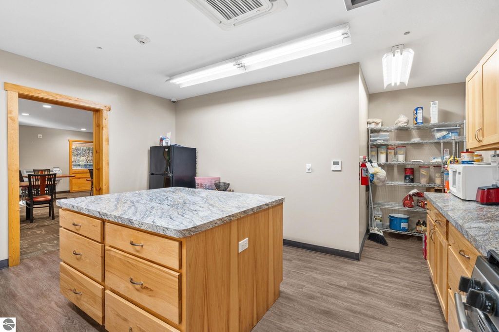 Kitchen interior featuring a central island with wooden drawers, granite countertop, and shelving stocked with kitchen supplies, adjacent dining area visible through an open doorway, suitable for a motel or resort property.