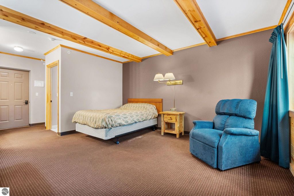Updated motel room with a queen bed, blue recliner, wooden side table, and natural light from a window, showcasing the inviting interior of a resort property near Torch Lake in Rapid City, MI.