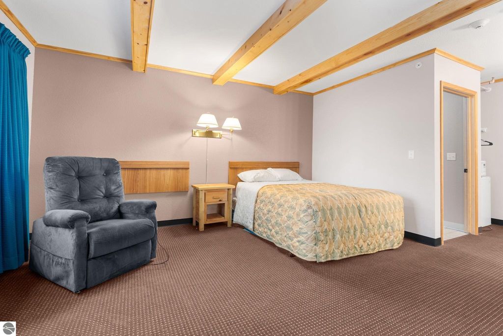 Updated motel room featuring a queen bed with patterned bedding, a comfortable gray recliner, wooden nightstand, and blue curtains, showcasing modern accommodations for guests near Torch Lake.