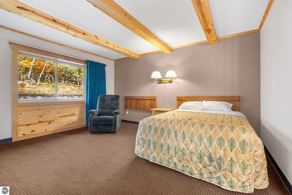Interior view of a motel room featuring a bed with patterned bedding, a bedside table, a blue armchair, and large windows with blue curtains, highlighting the updated accommodations for guests near Torch Lake.