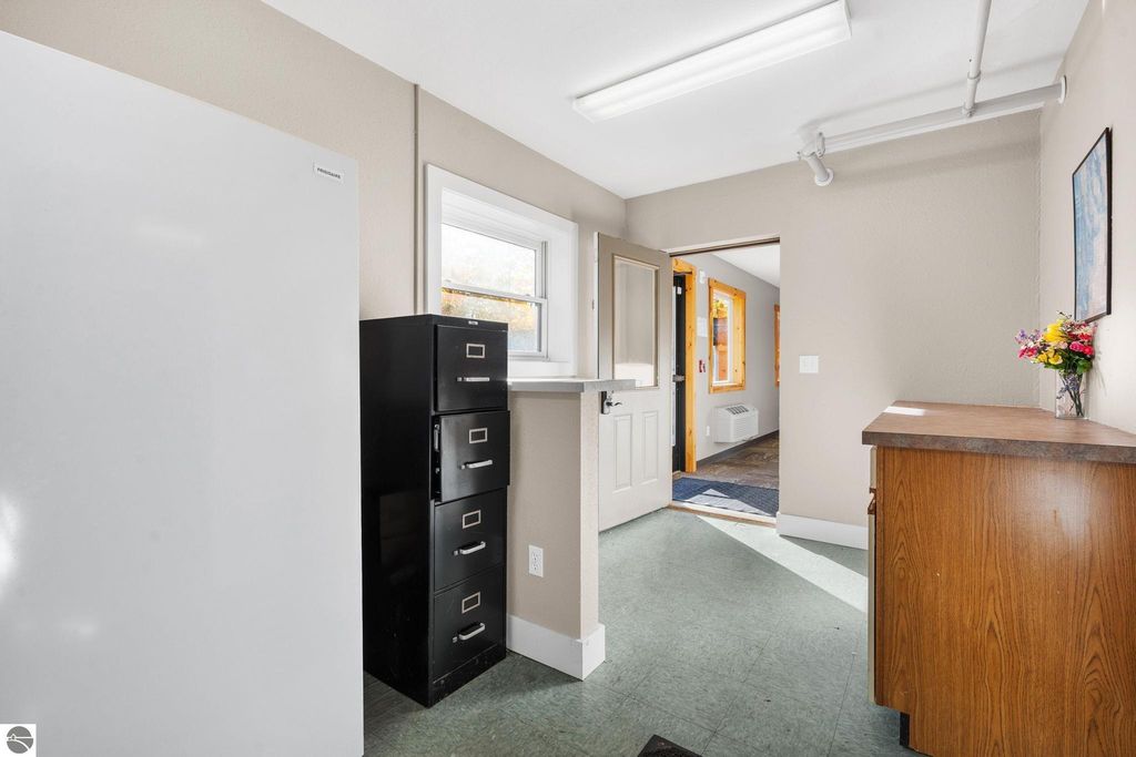 Interior view of a reception area featuring a white refrigerator, a black filing cabinet, and a wooden counter, with a doorway leading to a hallway; suitable for a motel or rental property.