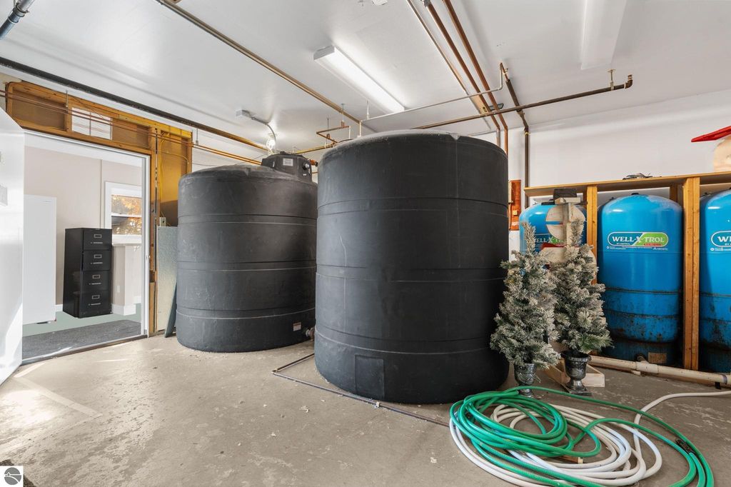 Interior view of a utility room featuring large black water tanks, blue pressure tanks labeled "WELLXTROL," green hoses, and a black filing cabinet, emphasizing the property's infrastructure for a motel and residential home near Torch Lake.