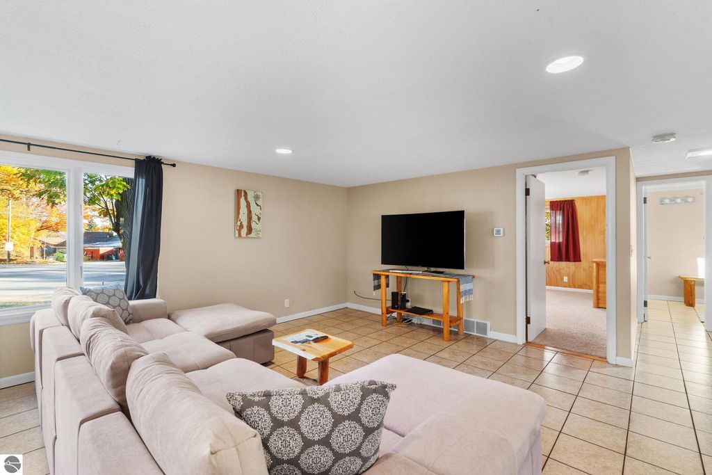 Living room of a resort property featuring a large sectional sofa, flat-screen TV on a wooden stand, and natural light from windows, emphasizing a welcoming atmosphere for guests near Torch Lake.
