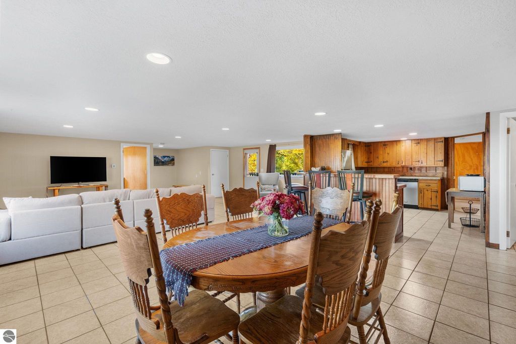 Interior view of a dining area with a wooden table and chairs, featuring a floral centerpiece, adjacent to a living space with a large sofa and television, showcasing a cozy and updated ambiance suitable for a resort property near Torch Lake, Michigan.