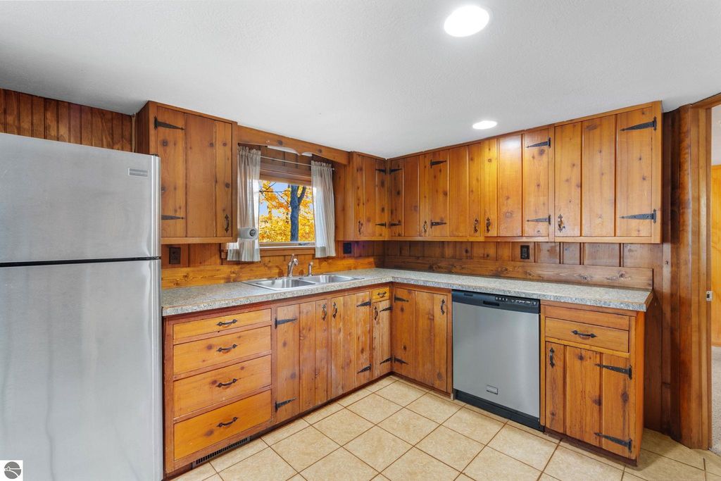 Updated kitchen with wooden cabinetry, stainless steel refrigerator, and double sink, featuring natural light from window overlooking trees, suitable for resort property at 12800 & 12862 Cherry Avenue, Rapid City, MI.