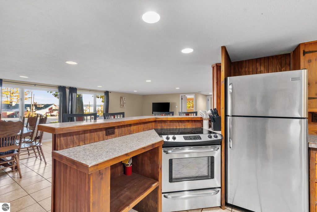 Modern kitchen interior with stainless steel appliances, including a stove and refrigerator, featuring a wooden bar counter and dining area, with large windows showcasing views of the outdoors, relevant to the resort property listing at 12800 & 12862 Cherry Avenue, Rapid City, MI.