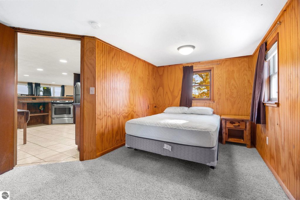 Interior view of updated motel room featuring a queen bed, wooden paneling, carpet flooring, and a connecting doorway to a kitchen area, highlighting the property’s potential for multi-room suite configurations at 12800 & 12862 Cherry Avenue, Rapid City, MI.