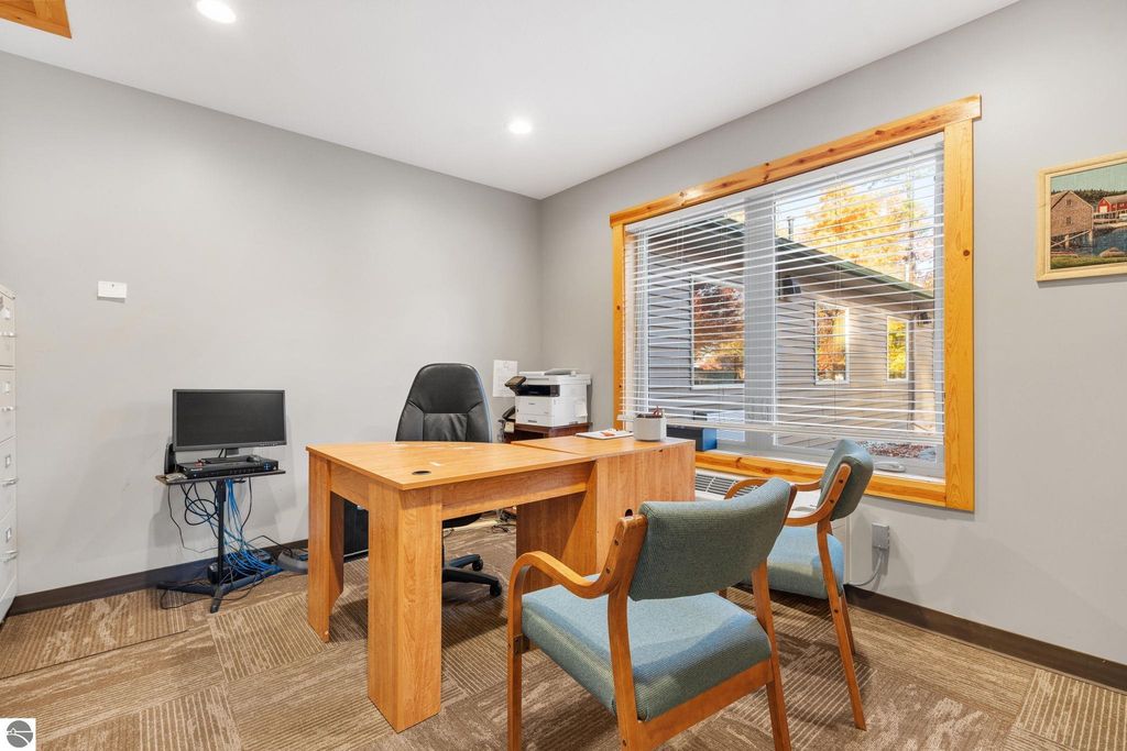 Office space featuring a wooden desk with two chairs, a computer monitor, and a printer, illuminated by natural light from a large window with blinds, located in a property near Torch Lake, suitable for management or administrative tasks.