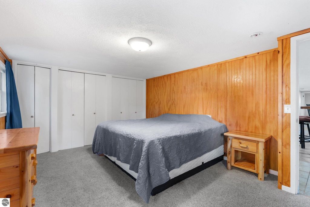 Bedroom featuring a queen-sized bed with gray bedding, wooden nightstand, and dresser, complemented by white closet doors and wood-paneled walls, ideal for a resort property near Torch Lake.