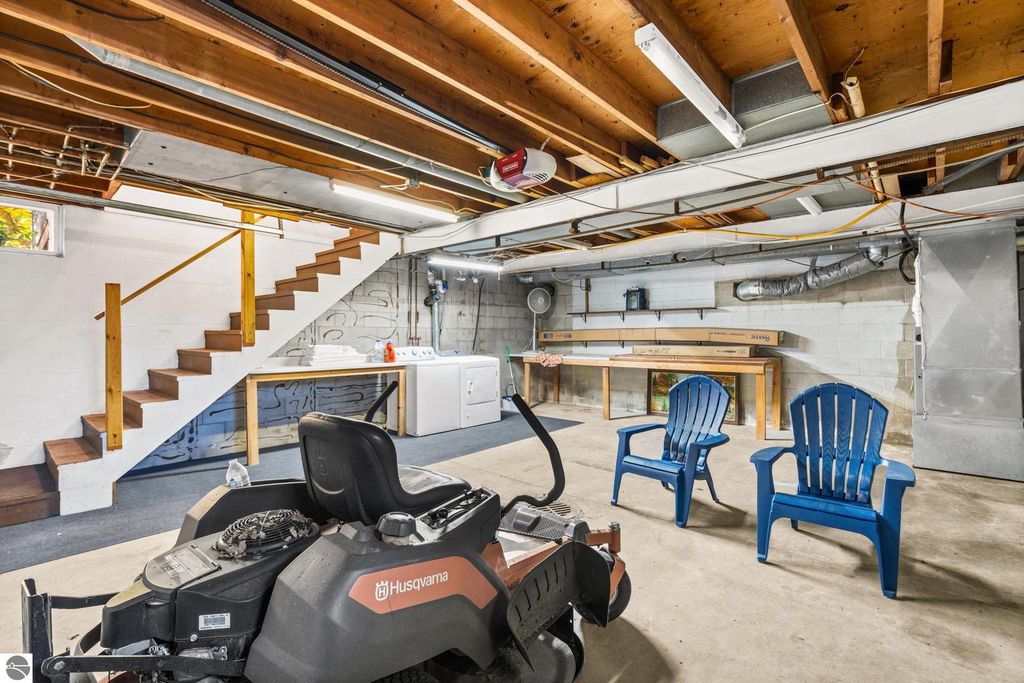 Basement interior featuring a Husqvarna lawn mower, blue Adirondack chairs, wooden stairs leading to the upper level, and a laundry area with a washing machine, highlighting functional space in a multi-family property for sale in Rapid City, MI.