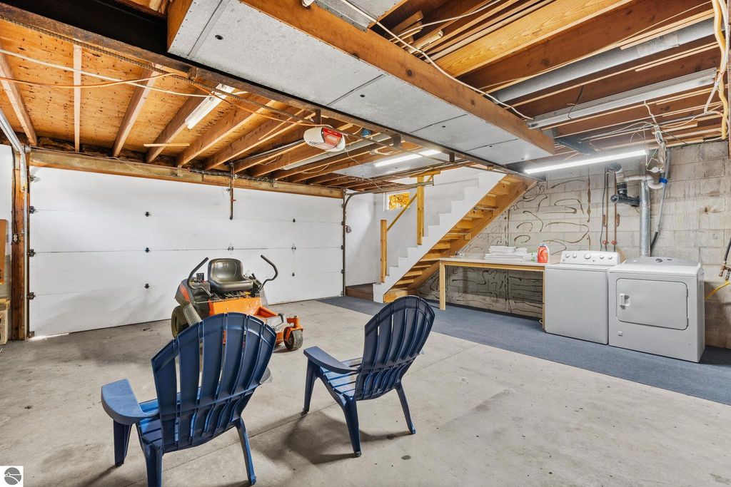 Basement laundry area featuring two blue chairs, a lawn mower, washing machine, and dryer, with a staircase in the background, suitable for property listing at 12800 & 12862 Cherry Avenue, Rapid City, MI.