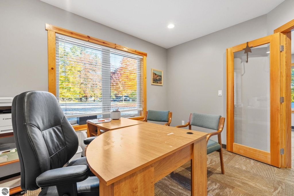 Office space featuring a wooden desk, ergonomic chair, and two additional chairs, with large windows showcasing autumn foliage, suitable for a property listing at 12800 & 12862 Cherry Avenue, Rapid City, MI.