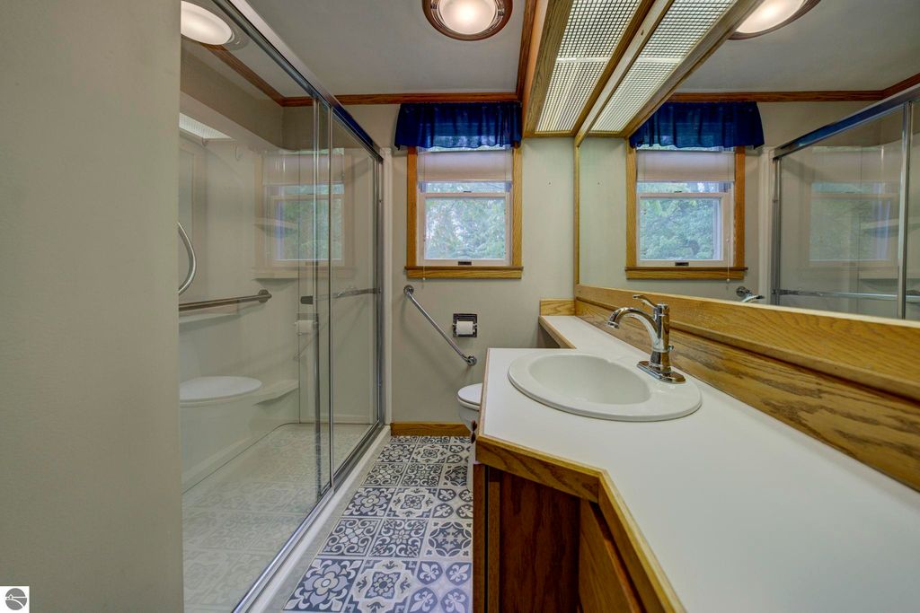 Bathroom with glass shower, decorative tiled floor, wooden cabinetry, and natural light from two windows.