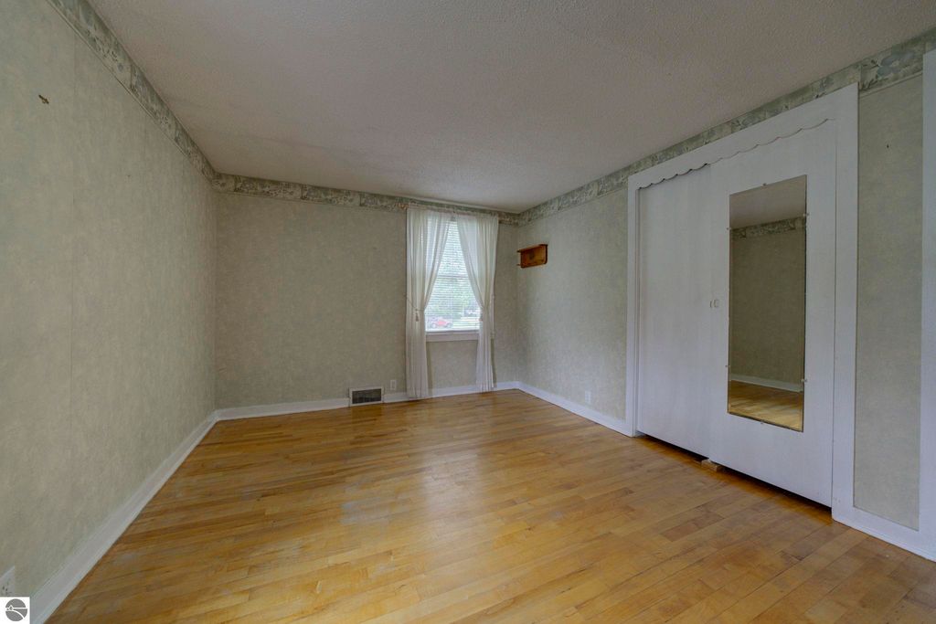 Bright and airy room with hardwood flooring, window draped with sheer curtains, and built-in closet with mirror, showcasing the inviting interior of the home at 624 Fenton Street, Kingsley, MI.
