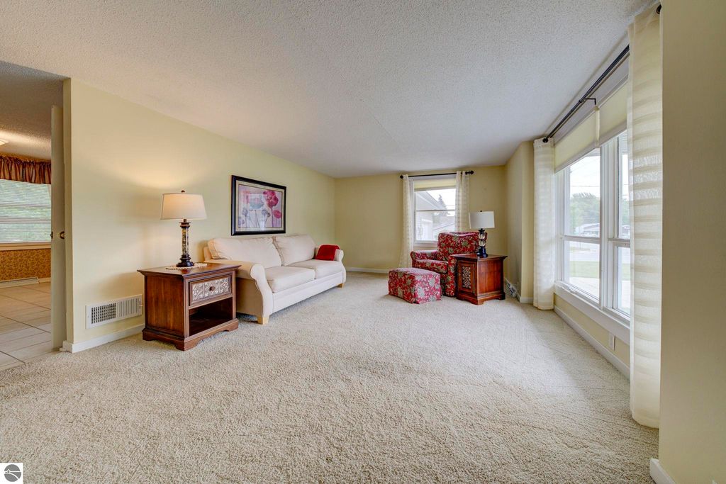 Bright and inviting living room with beige walls, plush carpet, a white sofa, floral accent chair, wooden side table with lamp, and large windows allowing natural light, showcasing the cozy atmosphere of the home at 624 Fenton Street, Kingsley, MI.